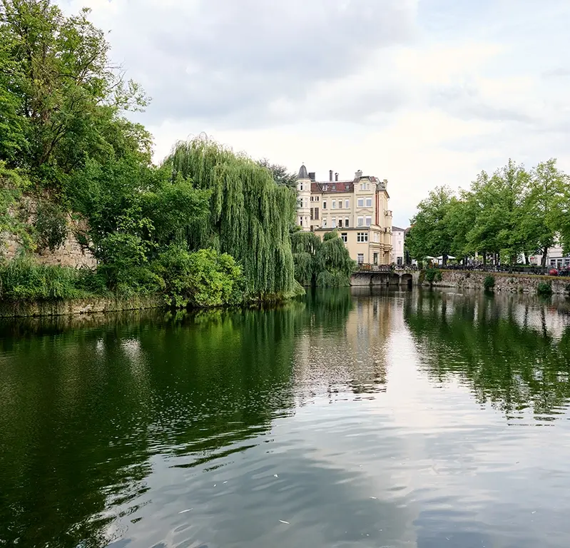 Landschaftsaufnahme von Detmold. Im Vordergrund ist ein Fluss, im Hintergrund ein schlossähnliches Gebäude.
