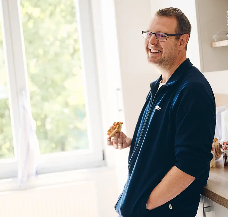 Mitarbeiter Herr Richert im Pausenraum. Er lehnt an einer Küchenzeile und unterhält sich mit einer weiteren Mitarbeiterin.