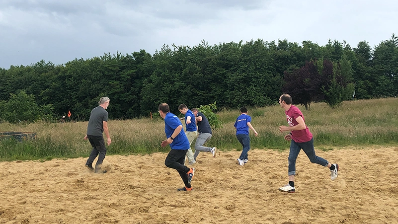 Ein Bild von mehreren Mitarbeiterinnen und Mitarbeitern, die im Sand Fußball spielen.